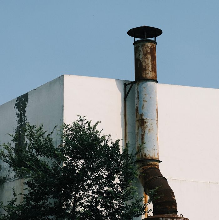 An urban scene featuring a rusty pipe on the corner of a white building with surrounding trees.